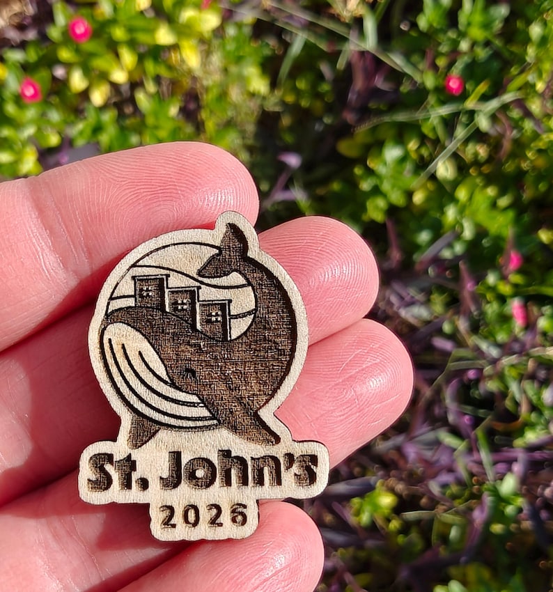 May include: Wooden emblem featuring a whale with buildings in its body, and the text "St. John's 2026". The emblem is made of light-colored wood with a darker, engraved design. The whale's tail curves around the buildings.