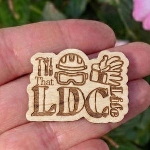 May include: A wooden laser-cut badge with the text "That LDC Life" and construction-themed icons. The badge features a hammer, hard hat, safety glasses, and work gloves. The badge is held in a hand.