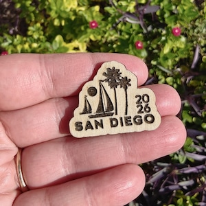 May include: A wooden souvenir featuring a sailboat, palm trees, and the text "SAN DIEGO 2026". The design is etched into the light-colored wood, with a sun and sailboat silhouette.
