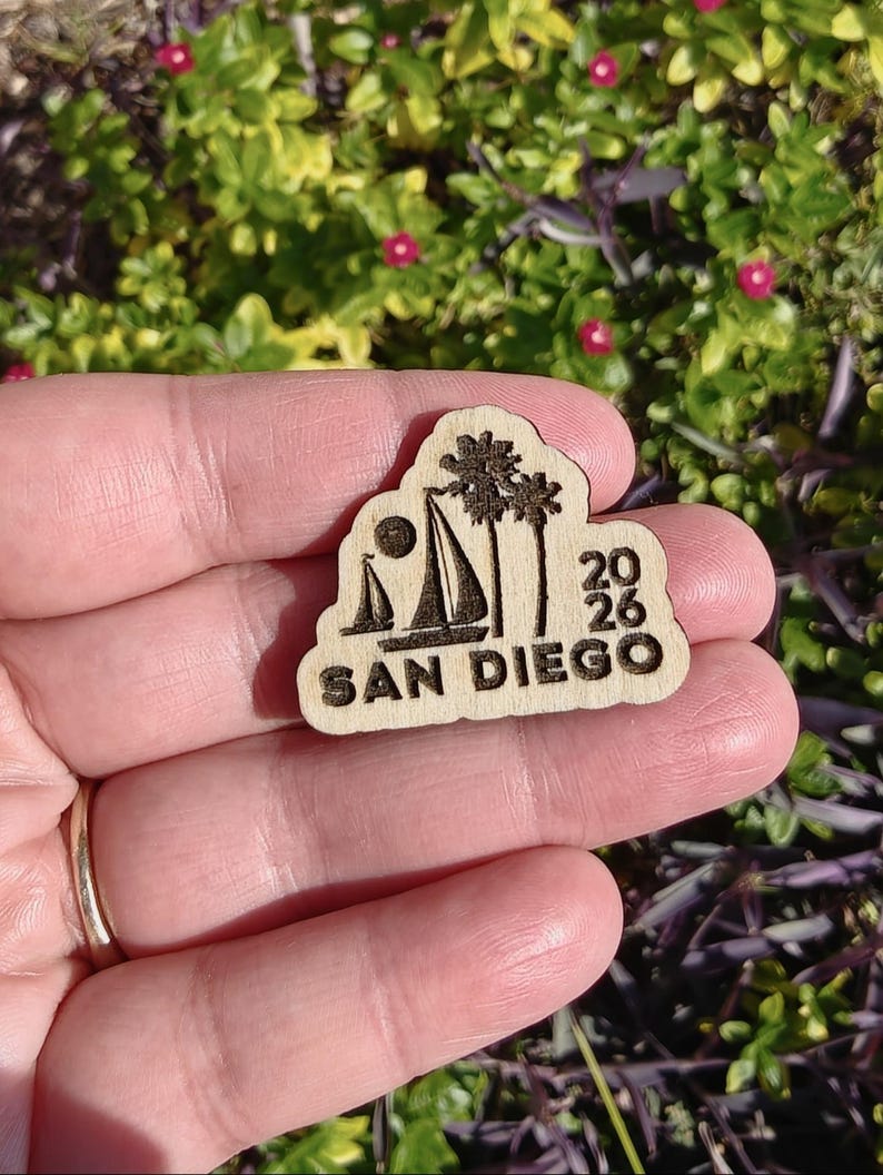 May include: A wooden souvenir featuring a sailboat, palm trees, and the text "SAN DIEGO 2026". The design is etched into the light-colored wood, with a sun and sailboat silhouette.