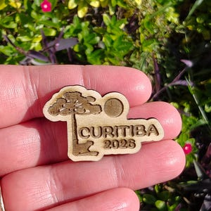 May include: A wooden Curitiba 2026 souvenir pin. The pin features a stylized tree, a sun, and the city name and year. The pin is held in a person's hand against a background of green foliage.