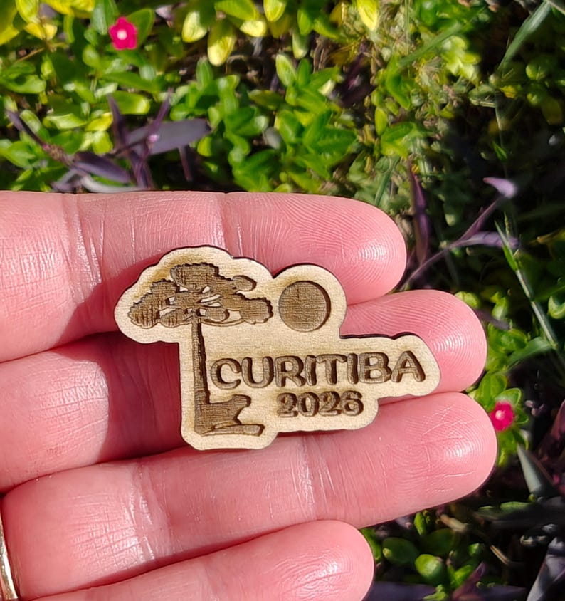 May include: A wooden Curitiba 2026 souvenir pin. The pin features a stylized tree, a sun, and the city name and year. The pin is held in a person's hand against a background of green foliage.