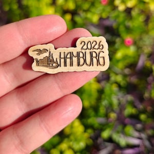 May include: A small, wooden souvenir featuring the text "2026 HAMBURG" in a stylized font. The design includes an illustration of a building and a bird. The item is held against a blurred green and pink background.