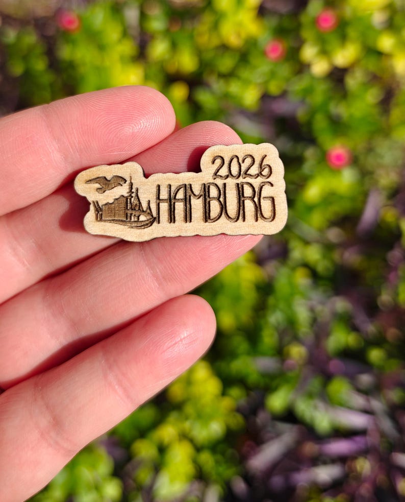 May include: A small, wooden souvenir featuring the text "2026 HAMBURG" in a stylized font. The design includes an illustration of a building and a bird. The item is held against a blurred green and pink background.