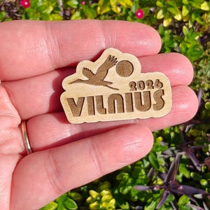 May include: A wooden souvenir featuring a stork in flight, a sun, and the text "VILNIUS 2026". The design is laser-cut from light-colored wood. The souvenir is held in a hand against a background of green foliage and small red flowers.