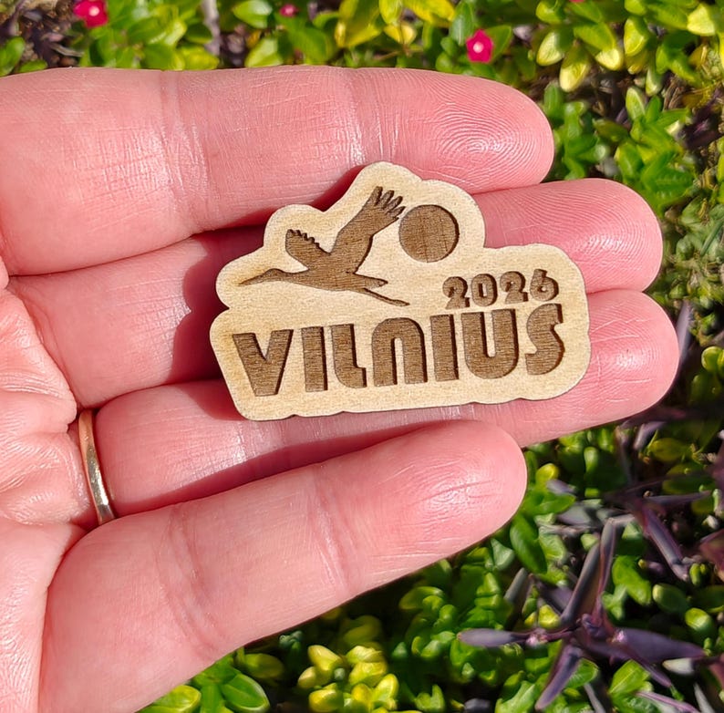 May include: A wooden souvenir featuring a stork in flight, a sun, and the text "VILNIUS 2026". The design is laser-cut from light-colored wood. The souvenir is held in a hand against a background of green foliage and small red flowers.