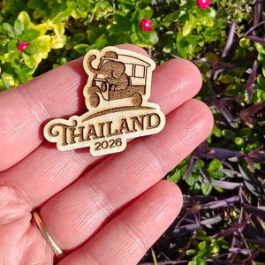 May include: A wooden souvenir featuring an elephant driving a tuk-tuk, with the word "Thailand" and the year "2026" engraved below. The design is in a light brown color, with a detailed illustration of the vehicle and elephant.