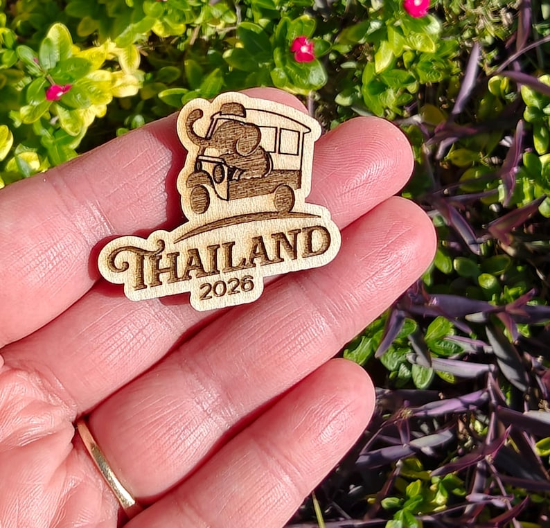 May include: A wooden souvenir featuring an elephant driving a tuk-tuk, with the word "Thailand" and the year "2026" engraved below. The design is in a light brown color, with a detailed illustration of the vehicle and elephant.