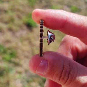 May include: A small, brown wooden earring with a vertical, ridged design. The earring post and backing are silver-colored. The earring is held between two fingers against a blurred, natural background.