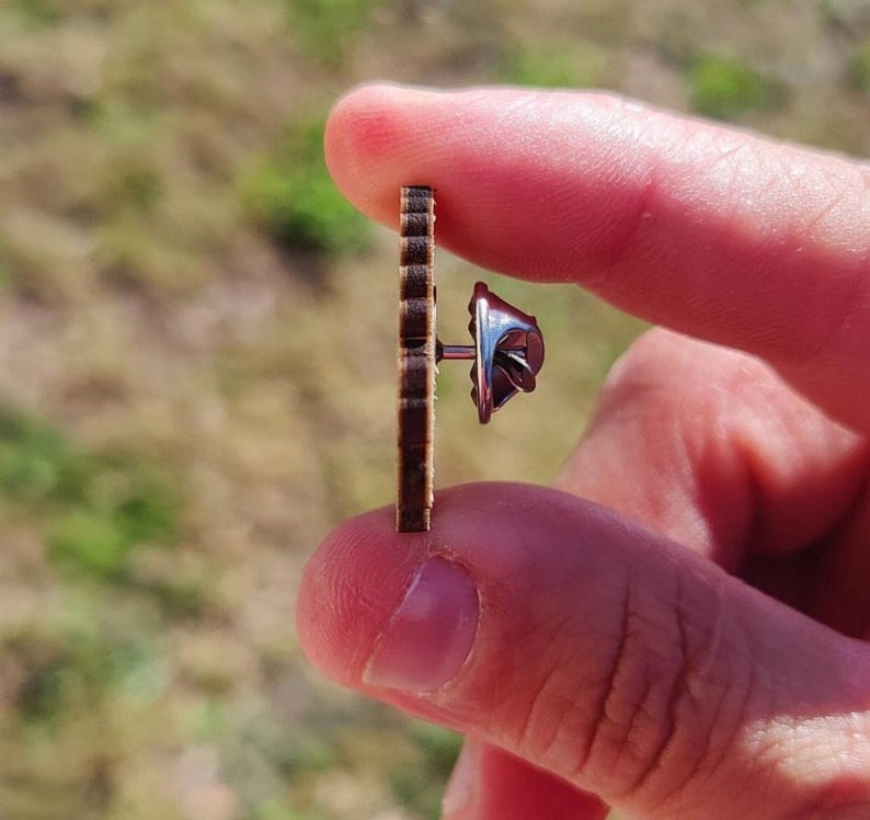 May include: A small, brown wooden earring with a vertical, ridged design. The earring post and backing are silver-colored. The earring is held between two fingers against a blurred, natural background.