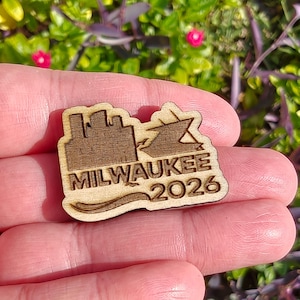 May include: A wooden pin featuring the city skyline, a ship, and the text "MILWAUKEE 2026". The pin is light brown and has a wave design at the bottom. The pin is held in a person's hand.