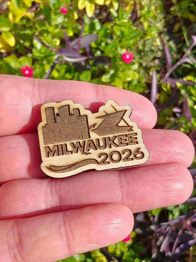 May include: A wooden pin featuring the city skyline, a ship, and the text "MILWAUKEE 2026". The pin is light brown and has a wave design at the bottom. The pin is held in a person's hand.