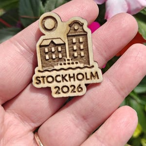 May include: A wooden pin featuring a laser-cut design of two buildings, a sun, and the text "STOCKHOLM 2026". The pin is held in a hand against a backdrop of colorful flowers.