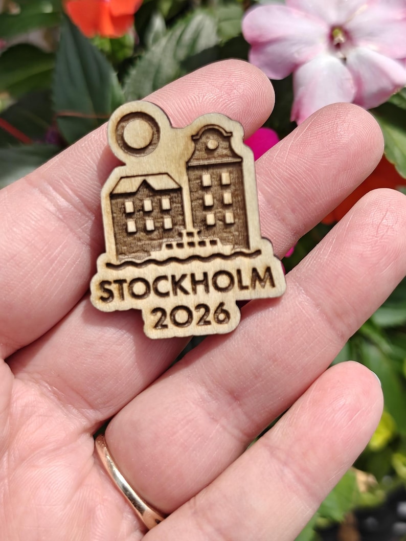 May include: A wooden pin featuring a laser-cut design of two buildings, a sun, and the text "STOCKHOLM 2026". The pin is held in a hand against a backdrop of colorful flowers.