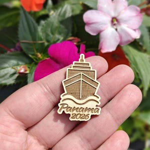 May include: A wooden ship-shaped pin with the text "Panama 2026" engraved on it. The pin is light brown and features a detailed ship design with waves at the bottom. The background includes colorful flowers.