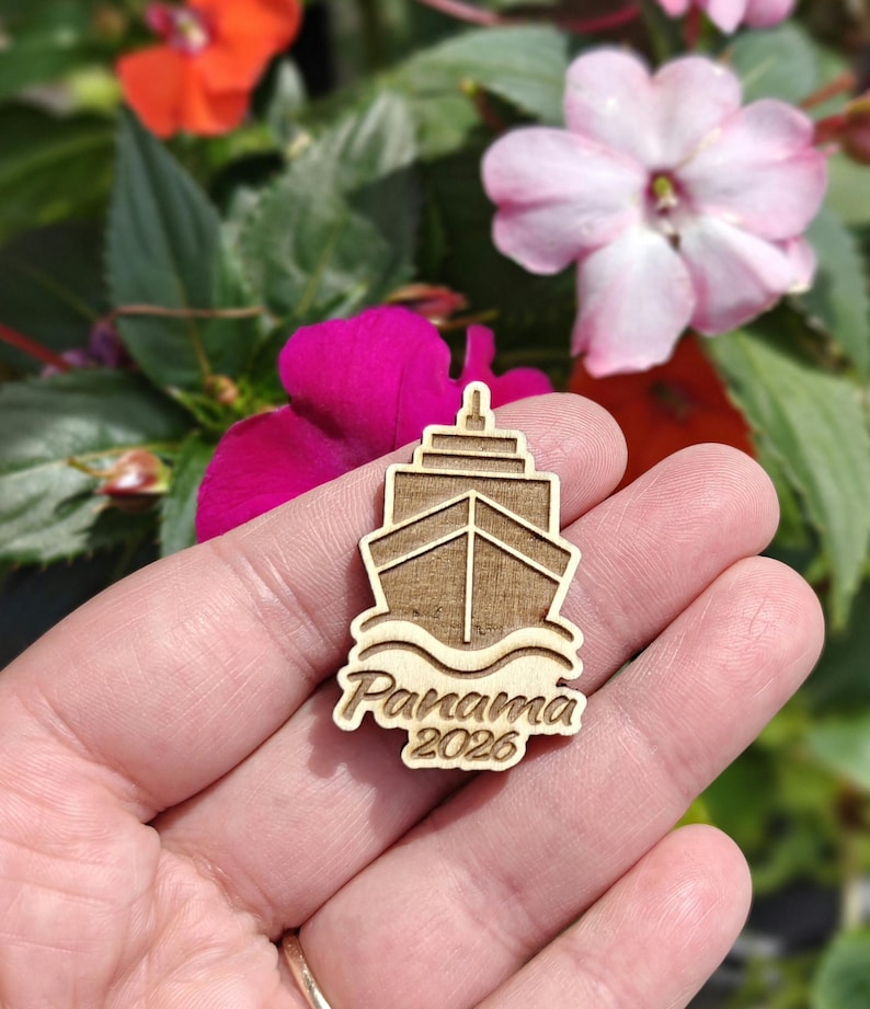 May include: A wooden ship-shaped pin with the text "Panama 2026" engraved on it. The pin is light brown and features a detailed ship design with waves at the bottom. The background includes colorful flowers.