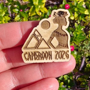 May include: A wooden laser-cut pin featuring a silhouette of a woman with a headdress, mountains, and a sun. The text "CAMEROON 2026" is at the bottom. The pin is held in a hand against a background of green leaves and pink flowers.