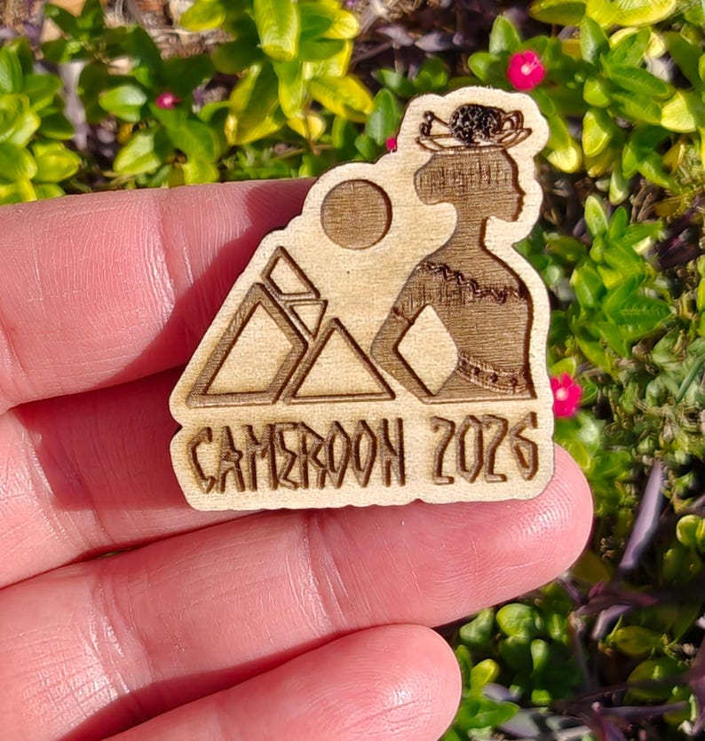 May include: A wooden laser-cut pin featuring a silhouette of a woman with a headdress, mountains, and a sun. The text "CAMEROON 2026" is at the bottom. The pin is held in a hand against a background of green leaves and pink flowers.
