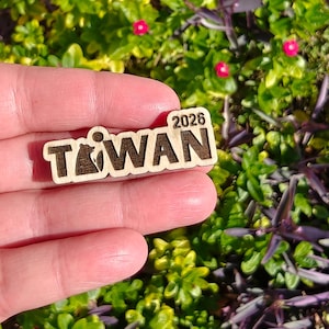 May include: A wooden pin with the word "TAIWAN" and the year "2026" engraved on it. The pin features a bear graphic within the letter "T". The pin is held in a hand against a background of green foliage and small pink flowers.