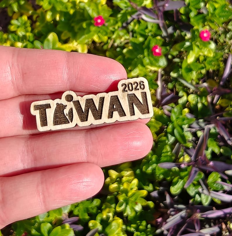 May include: A wooden pin with the word "TAIWAN" and the year "2026" engraved on it. The pin features a bear graphic within the letter "T". The pin is held in a hand against a background of green foliage and small pink flowers.