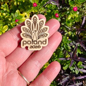 May include: A wooden pin featuring a peacock design with the word "Poland" and the year "2026" engraved. The pin is held in a hand against a backdrop of green foliage and small pink flowers.