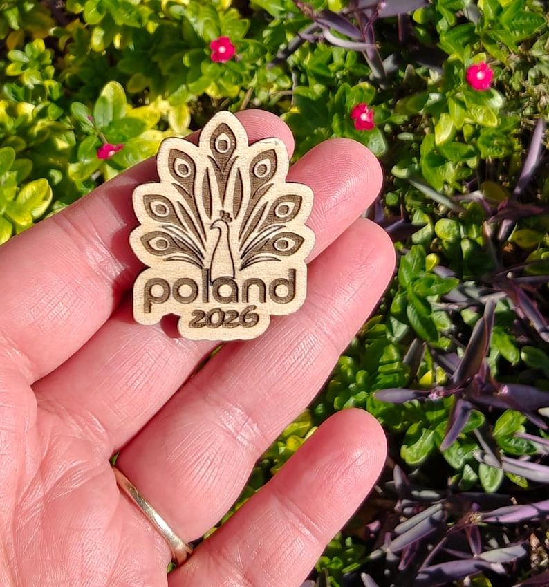 May include: A wooden pin featuring a peacock design with the word "Poland" and the year "2026" engraved. The pin is held in a hand against a backdrop of green foliage and small pink flowers.
