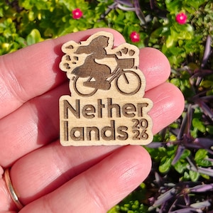 May include: Wooden pin featuring a silhouette of a person riding a bicycle with a basket, with the text "Netherlands 2023". The pin is made of light brown wood and has a detailed design.