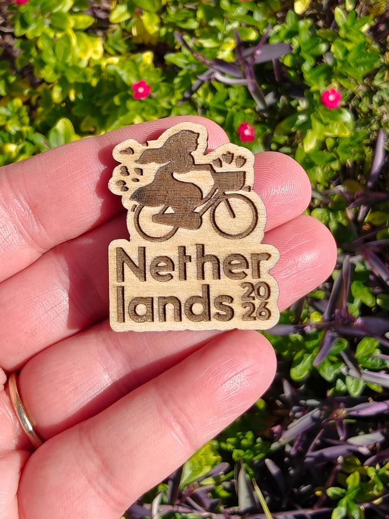 May include: Wooden pin featuring a silhouette of a person riding a bicycle with a basket, with the text "Netherlands 2023". The pin is made of light brown wood and has a detailed design.