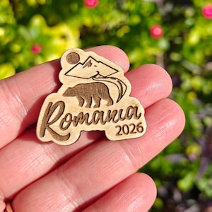 May include: Wooden Romania souvenir with a bear, mountains, and the year 2026. The design is laser-cut and features the word "Romania" in a stylized font. The background is blurred, with green and pink tones.