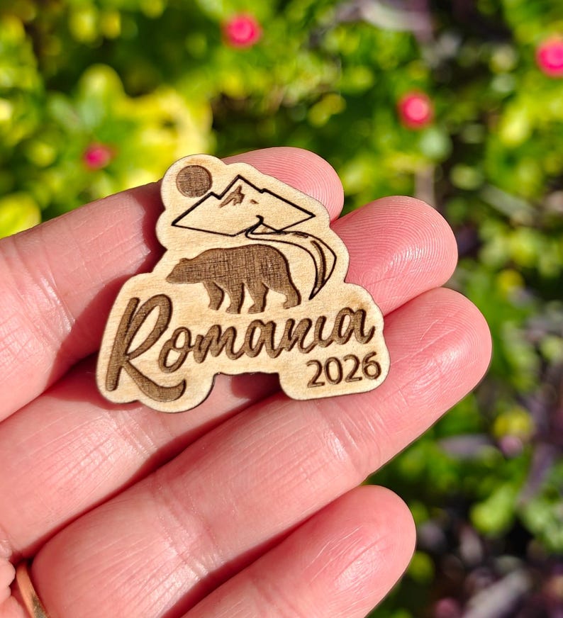 May include: Wooden Romania souvenir with a bear, mountains, and the year 2026. The design is laser-cut and features the word "Romania" in a stylized font. The background is blurred, with green and pink tones.
