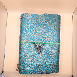 May include: A teal leather journal with embossed floral patterns and gold accents. The journal is secured with a green elastic band and a silver Celtic knot charm. Silver metal clasps secure the journal's cover.
