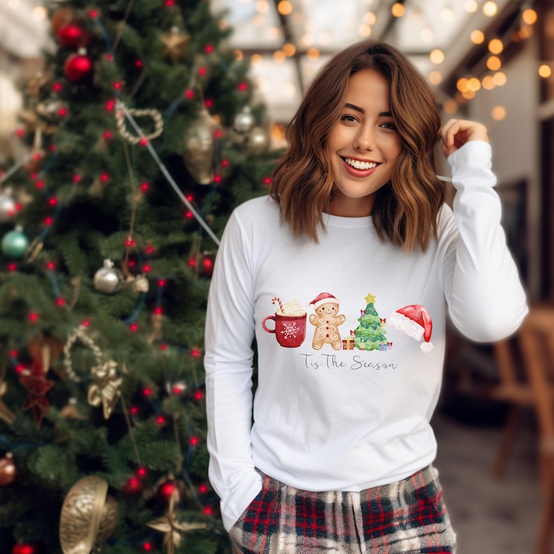 Puede incluir: Camiseta blanca de manga larga con un diseño navideño. El diseño incluye una taza roja, un hombre de jengibre, un árbol de Navidad y un gorro de Papá Noel. El texto "Tis The Season" está debajo.