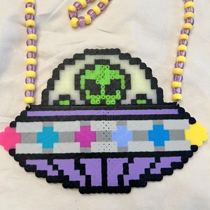 Peut inclure: Un OVNI pixelisé violet et blanc avec un extraterrestre vert à l'intérieur. L'OVNI est attaché à un collier de perles jaunes et violettes.