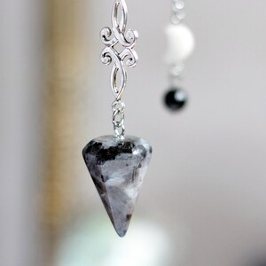 Op de afbeelding: Een pendel met een facetgeslepen, grijszwarte steen die aan een zilveren ketting hangt. De ketting heeft een sierlijk, decoratief schakel. Een tweede ketting met een witte en zwarte kraal is op de achtergrond zichtbaar.