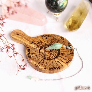 Puede incluir: Un tablero de péndulo de madera con un péndulo de cristal verde y una cadena plateada. El tablero tiene las palabras "Sí", "No", "Tal vez" y "Reformular" inscritas en él. El tablero está rodeado de cristales.