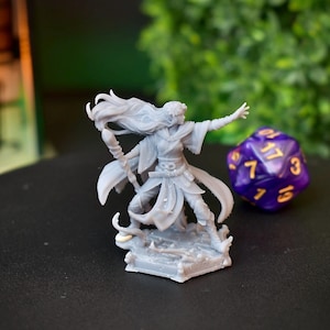 Könnte beinhalten: Eine graue 3D-gedruckte Miniatur einer weiblichen Figur mit langen, fließenden Haaren, die eine Robe trägt und einen Stab hält. Die Figur steht auf einem sechseckigen Sockel mit aufwendigen Details. Ein lila und goldener Würfel befindet sich im Hintergrund.