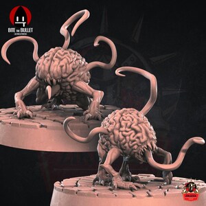 Op de afbeelding: Twee 3D-geprinte miniatuurfiguren van hersenenachtige wezens met meerdere tentakels. De figuren zijn geschilderd in een vleeskleurige roze kleur en staan op ronde bases.