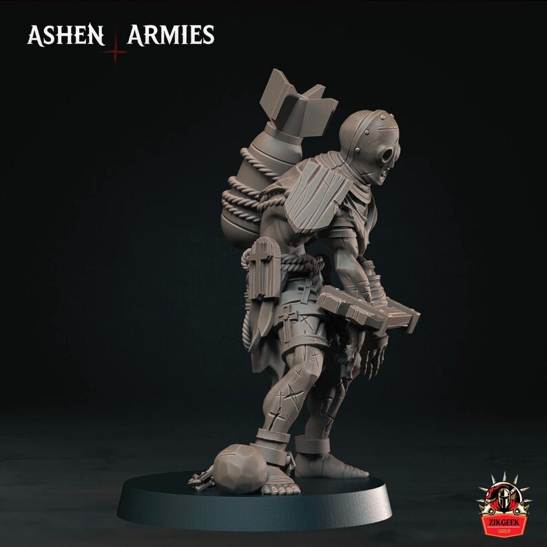 May include: A grey miniature figurine of a humanoid character with a large bomb strapped to its back with rope. The figure wears a helmet, shoulder armor, and shorts with cross motifs, holding a rectangular object while standing on a round base.