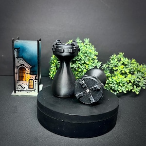 Op de afbeelding: Zwarte flessenstoppers in de vorm van een schaaktoren. Eén stopper staat rechtop, de andere ligt op zijn kant. De stoppers staan op een zwart oppervlak met groen gebladerte op de achtergrond. Een decoratieve huisvormige lamp staat links.