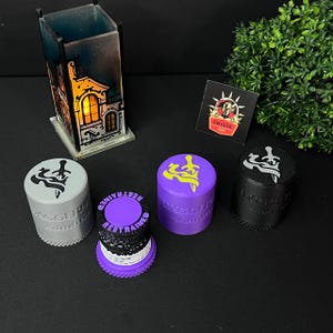 May include: An assortment of cylindrical grinders in various colors, including gray, purple, and black. Each grinder features a unique design on the top and the text "DAGGER" and "RESTRAINED" on the sides. A decorative lantern and a sticker are also visible.
