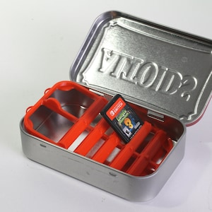 May include: A silver metal tin with a red plastic insert for storing Nintendo Switch game cartridges. The lid of the tin has the word "VOID?" embossed on it.