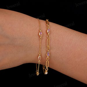 May include: Two gold bracelets are displayed against a dark background. One bracelet features oval-shaped pink gemstones, while the other has a chain link design with similar gemstones. The bracelets are delicate and elegant.
