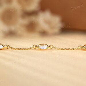 May include: A delicate gold chain bracelet featuring oval-shaped, clear gemstones. The bracelet's links are small and connected, with the gemstones evenly spaced along the chain. The background is a soft, warm color, highlighting the jewelry.
