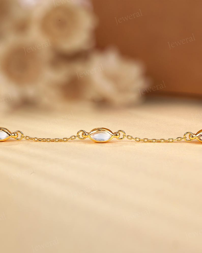 May include: A delicate gold chain bracelet featuring oval-shaped, clear gemstones. The bracelet's links are small and connected, with the gemstones evenly spaced along the chain. The background is a soft, warm color, highlighting the jewelry.