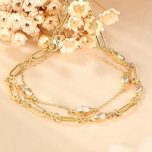 May include: A gold-toned necklace with two strands, featuring oval-shaped clear stones and elongated link chain details. The necklace is displayed with a backdrop of white flowers, creating an elegant and feminine aesthetic.