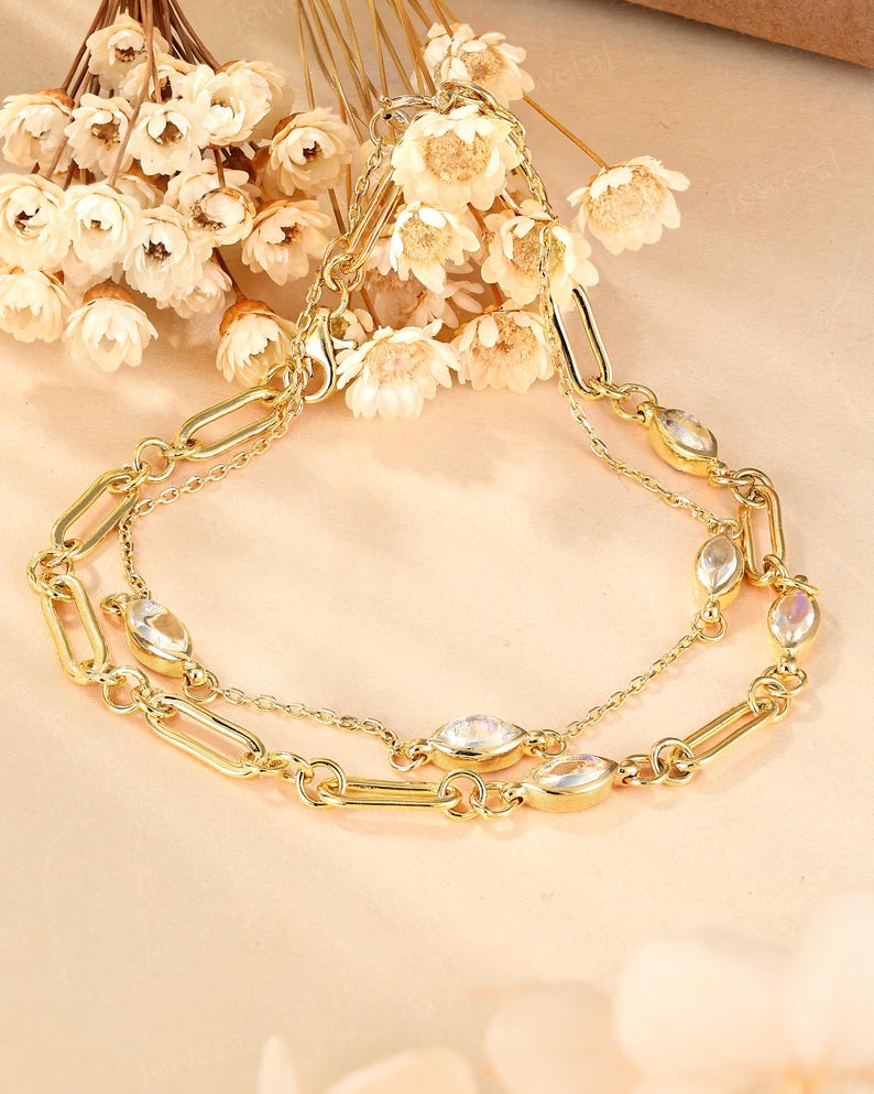 May include: A gold-toned necklace with two strands, featuring oval-shaped clear stones and elongated link chain details. The necklace is displayed with a backdrop of white flowers, creating an elegant and feminine aesthetic.