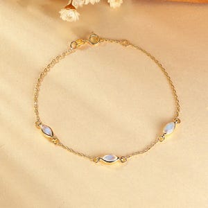 May include: A delicate gold bracelet featuring three oval-shaped, iridescent gemstones. The bracelet has a fine chain and a secure clasp. The gemstones have a soft, milky white color. The jewelry is displayed on a neutral background.