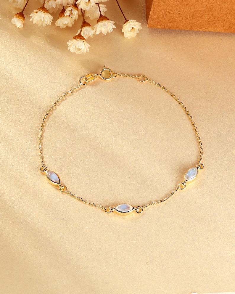 May include: A delicate gold bracelet featuring three oval-shaped, iridescent gemstones. The bracelet has a fine chain and a secure clasp. The gemstones have a soft, milky white color. The jewelry is displayed on a neutral background.