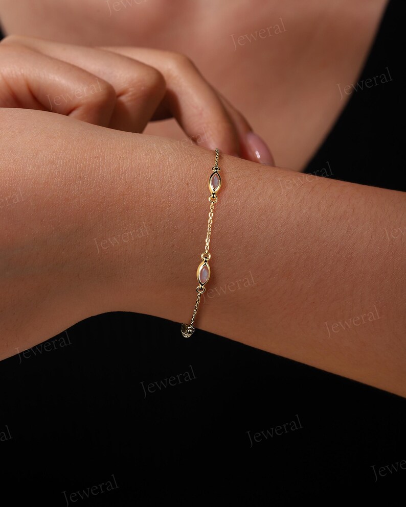 May include: A delicate gold bracelet with three oval, light pink gemstones. The bracelet is attached to a delicate chain and is worn on a wrist. The bracelet is a piece of jewelry.