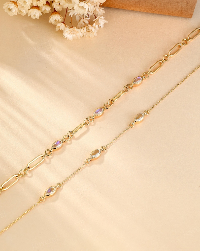 May include: Two gold-colored necklaces. One features oval-shaped links and iridescent stones, while the other has a delicate chain with similar stones. The necklaces are displayed on a light beige surface, with white flowers in the background.